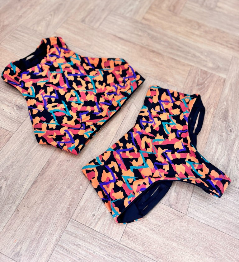 This image features the "Hitchhiker Bottoms- NOW & THEN" by Strange, modeled in a daring and playful spirit. The high-waisted bikini bottoms are showcased in a vibrant cantaloupe color, designed to reverse, as indicated, to a medina pattern.