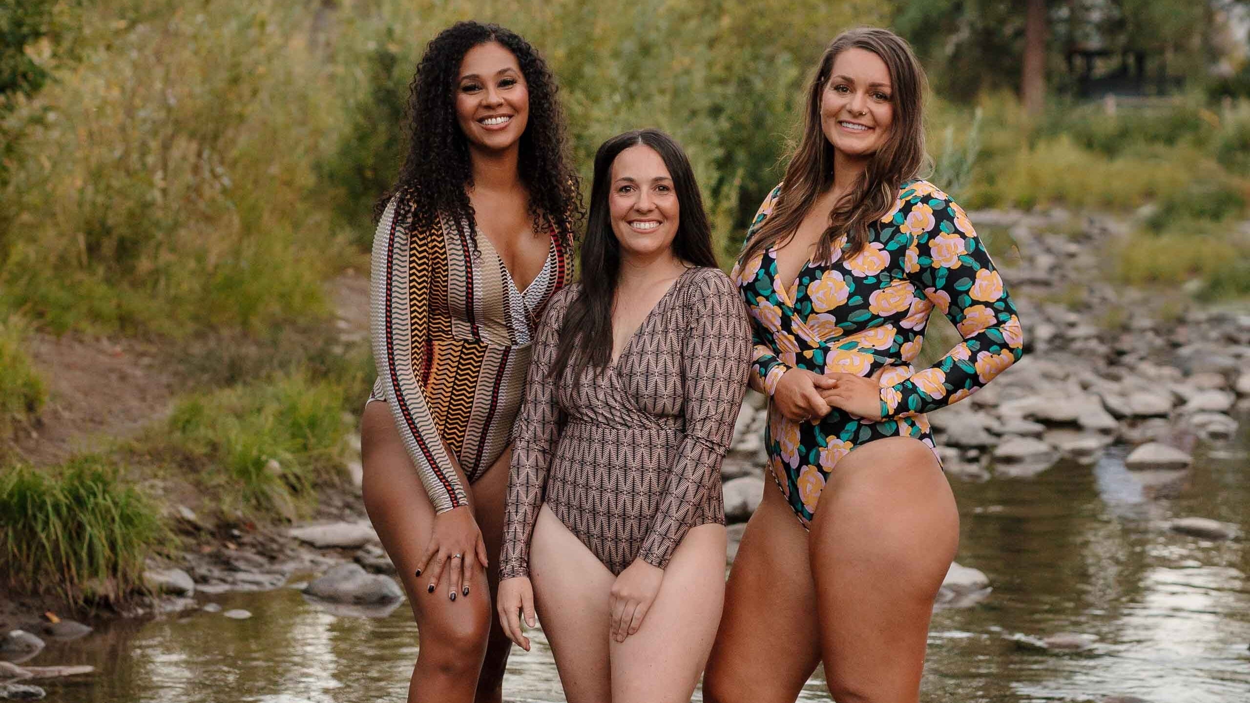 Three women standing in a natural setting with trees and water.
